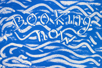 Booking now inscription on a blue colored sand with waves, top view, flat lay
