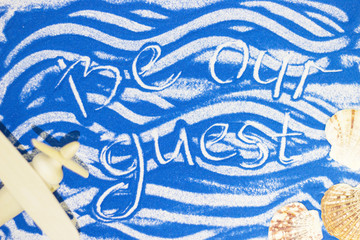 Be our guest inscription on a blue colored sand with waves, sea shells, toy airplane, top view, flat lay