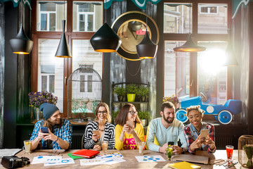 Multi ethnic group of young friends sitting with phones at the big table in the modern cafe interior