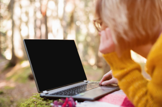 A mock-up image of a woman's hand using and typing on a laptop with a blank black desktop on a plaid grass lying in the forest. The concept of freelancing in freedom from a permanent place of work and