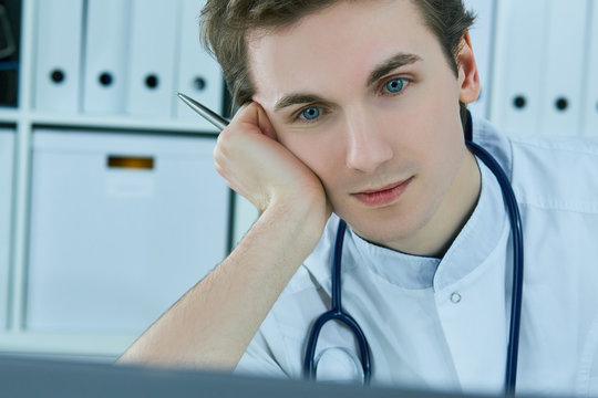 Close-up portrait of a young male doctor, propped his head by the hand working on his computer in medical office.