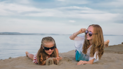 Two teenage are sitting on a sandy beach near the sea. Girl playing, talk to each other. Sisters are dressed in dresses and sunglasses. Children have real emotions of happiness.