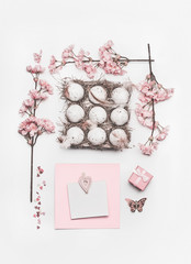 Beautiful pastel pink Easter layout with blossom decoration, hearts, eggs in carton box and greeting card mock up on white desk background, top view, flat lay.