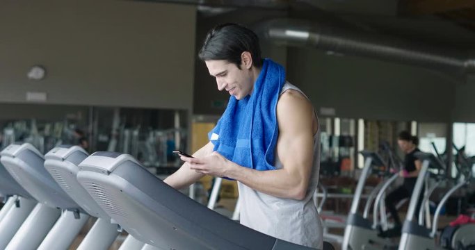 Handsome guy texting a message on is phone while he is walking on the treadmill, with the gym in the backround