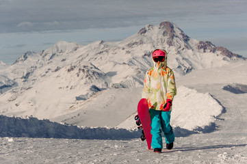 Female snowboarder in sportswear walking on the background of mountain peaks