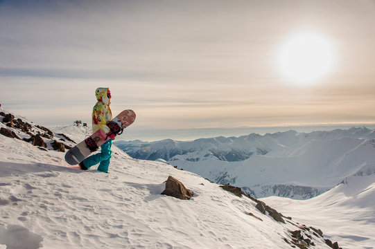 Female snowboarder in stylish sportswear walking on the background of the mountain peaks