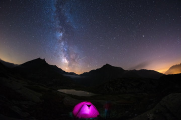 Fototapeta na wymiar Camping under starry sky and milky way at high altitude on the Alps. Illuminated tent in the foreground and majestic mountain peak in the background. Adventure and exploration in summertime.