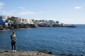 Young millennial female of Caucasian ethnicity holding a phone in the hand and walking away from the coast of Los Abrigos village, in Tenerife, Canary Islands, Spain