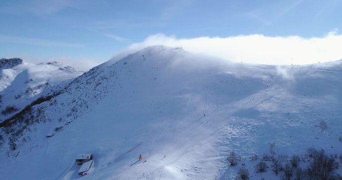 Forward aerial over winter snowy mountain top ski tracks resort with skier people skiing.Sunny day,foggy clouds.Fog rising.Alps mountains snow season establisher.4k drone flight sport establishing