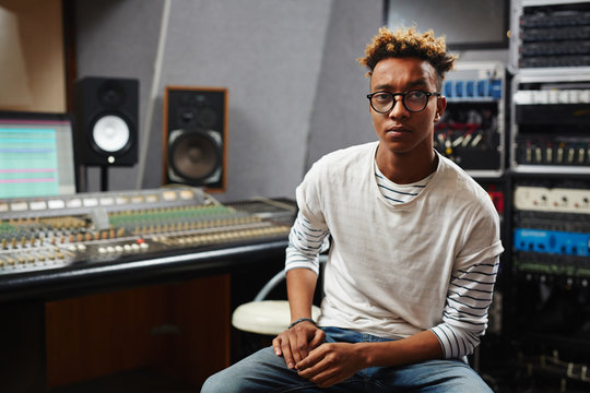 Young African-american deejay in eyeglasses looking at camera while sitting in studio of records