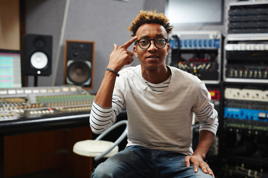 Young record studio staff in jeans, pullover and eyeglasses pointing at his temple and looking at camera