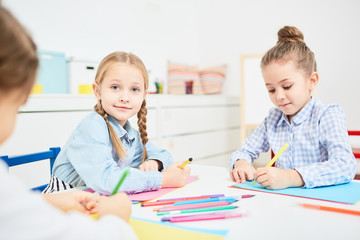 Adorable child with crayon looking at camera while drawing among her friends in kindergarten