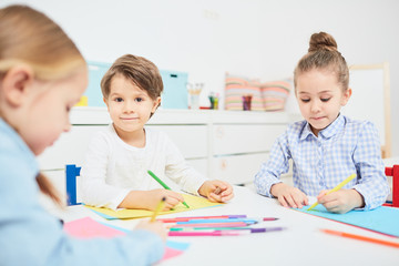Fototapeta premium Handsome little boy looking at camera while drawing with crayons between two girls