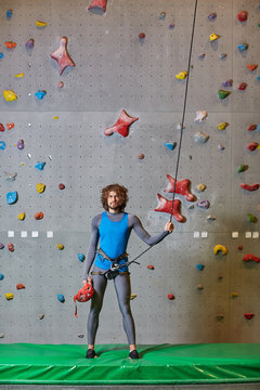 Active guy in blue sportive vest and grey leggins standing on mat and holding by strung climbing rope