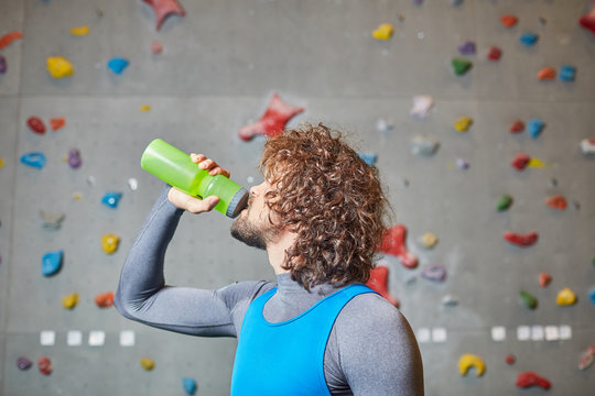 Curly young man drinking water from bottle on background of climbing wall