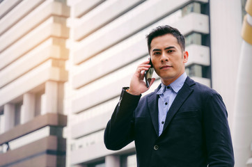 young smart asian business man wearing modern black suit making phone call with mobile smart phone in building city background, internet network connection, technology communication, financial concept