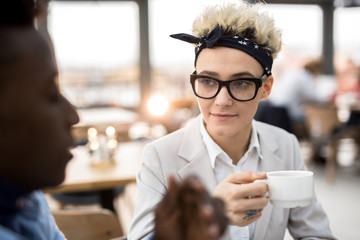 Serious woman in eyeglasses and headband having tea or coffee and listening to her colleague during conversation
