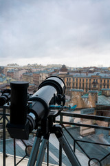 Black telescope on tripod in front of window with urban scene and grey sky outside