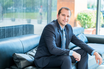 Portrait of a handsome mature businessman, relaxing on a couch in a business lounge, looking at the camera with a positive expression