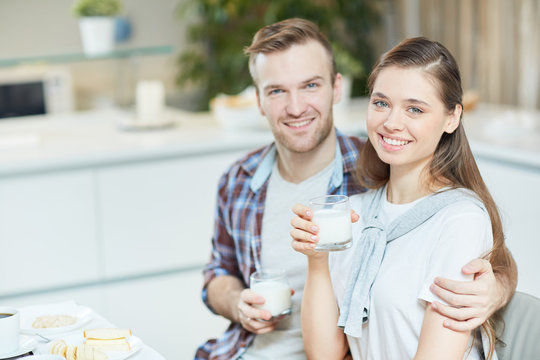 Happy woman and her husband with glasses of milk looking at camera while having breakfast