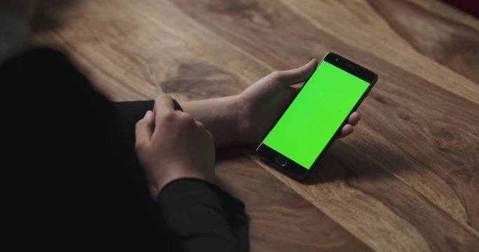 Slow motion handheld female teen girl using smartphone with green screen sitting at the table