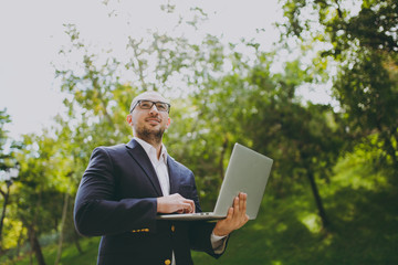 Young successful smart businessman in white shirt, classic suit, glasses. Man standing and working on laptop pc computer in city park outdoors on nature background. Mobile Office, business concept.