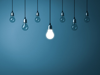 One hanging light bulb glowing and standing out from unlit incandescent bulbs on dark cyan background. 3D rendering.
