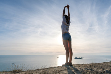 Unrecognizable woman in light blouse and denim shorts stretching on the hill with seascape Back view