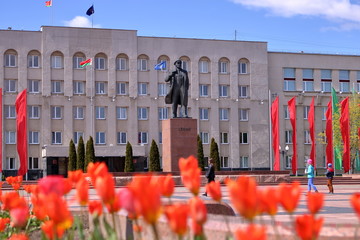 Pomnik Lenina na Białorusi, na pierwszym planie niewyraźne pomarańczowe tulipany, w tle budynek z wieloma oknami i flagą Białorusi, kilka osób na placu przed pomnikiem, na cokole cyrylicą LENIN