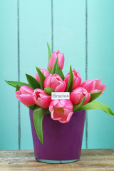 Gracias (thank you in Spanish) card with pink tulips in purple flower pot on blue wooden surface
