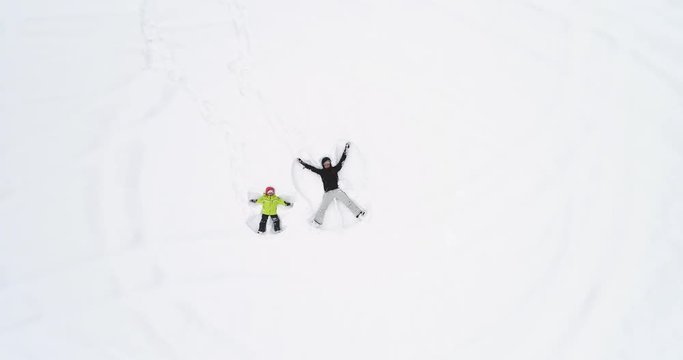 Overhead aerial top view over mother and child daughter lying doing snow angel on white covered winter snowy field.Mom and girl family people enjoy and have fun outdoors.4k straight-down perspective