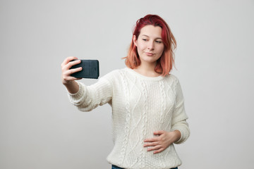 Naklejka premium Smiling young redhead woman with red hair making selfie using smart phone. Girl in white sweater taking photograph with mobile phone, looking with pretty smile at the camera of electronic device