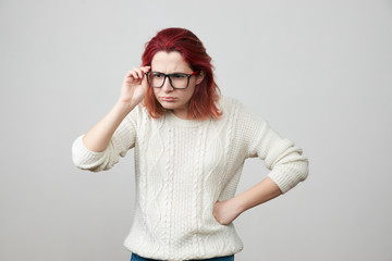 Portrait of young woman in white sweater with confused and gloomy expression, correcting glasses and puts one hand on her hip, isolated over gray background. The girl is indignant