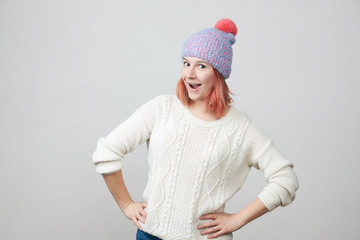 Young beautiful red-haired girl in knitted hat and sweater smiling looking at camera over grey background. 