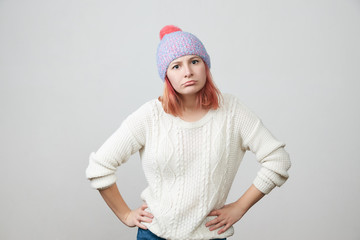 Portrait of a young beautiful red-haired girl in knitted hat and sweater indignantly and strictly looking at camera with her hands on her hips over grey background with copy space