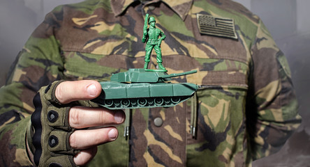 Photo of a military man in camouflage jacket holding a soldier toy standing on tank.