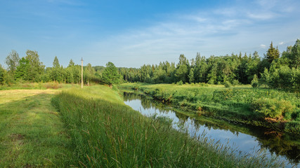 a picturesque summer landscape. A narrow river with a sloping meadow and dense forest on high coasts