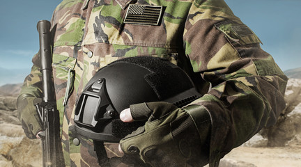 Photo of a soldier in military jacket holding helmet and standing on desert background.