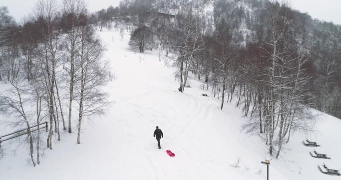 Aerial follow man father pulling red bobsled on snowy rise in winter.Dad bringing bobsleigh on snow.Family people enjoy together and have fun outdoors.4k drone flight video