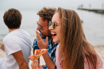 Funny attractive friends spending time with pleasure on the beach, cheerfully smiling, eating pizza.