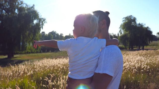 Young dad holding his child on hands at the meadow. Little son and handsome father pointing to something at the field. Happy family spending time together. Summer landscape at background. Close up