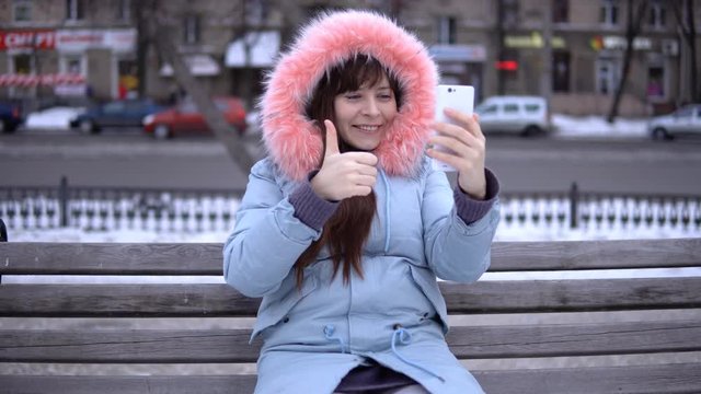 A young attractive woman in a gray coat makes a video call, she sits on a bench outside on a winter day.