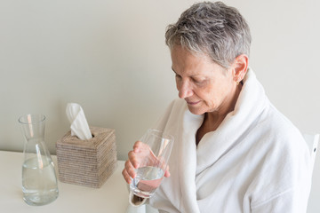 High angle view of older woman in white bathrobe drinking glass of water (selective focus)