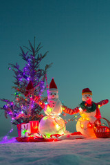 Two snowmen celebrate Christmas and New Year together near Christmas tree. Christmas tree decorated with Christmas toys and garlands on background. Snowmen wear warm clothes and wave their hands
