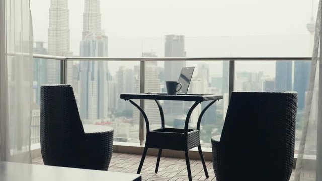 Teo chairs and a table with cup of coffee and open laptop at terrace with beautiful view over the city. Luxury apartment balcony and interior.