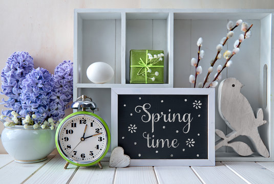 Old alarm clock, blue hyacinth flowers and a display cabinet with different objects. Chalk text "Springtime" on the blackboard