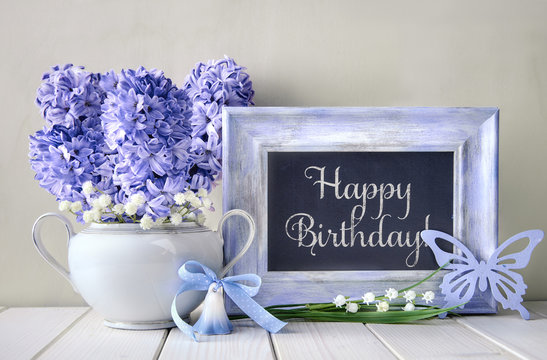 Blue decorations and hyacinth flowers on white table, blackboard with text "Happy Birthday"