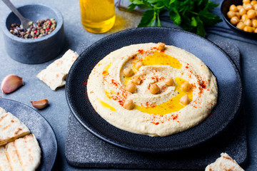 Hummus, chickpea dip, with spices and pita, flat bread in a black plate on grey stone background. Top view.