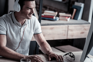 Favorite song. Successful adult entrepreneur looking at a screen of his computer with a cheerful smile on his face while sitting at a table and listening to music.