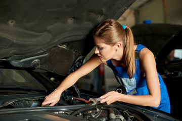 Young woman mechanics repairing or inspecting a car and holds a spanner in her hand. Girl is dressed in working clothes
